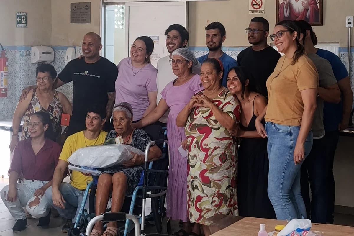 Voluntários e moradoras reunidos no refeitório da Casa de Nazaré, no Dia das Mães, com um presente embalado no colo de uma das senhoras.