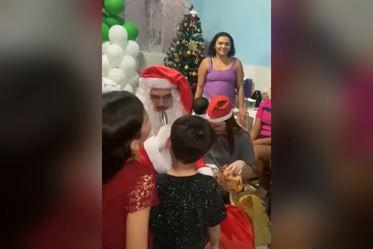 Papai Noel entregando presentes para crianças numa sala decorada com árvore de Natal e balões.